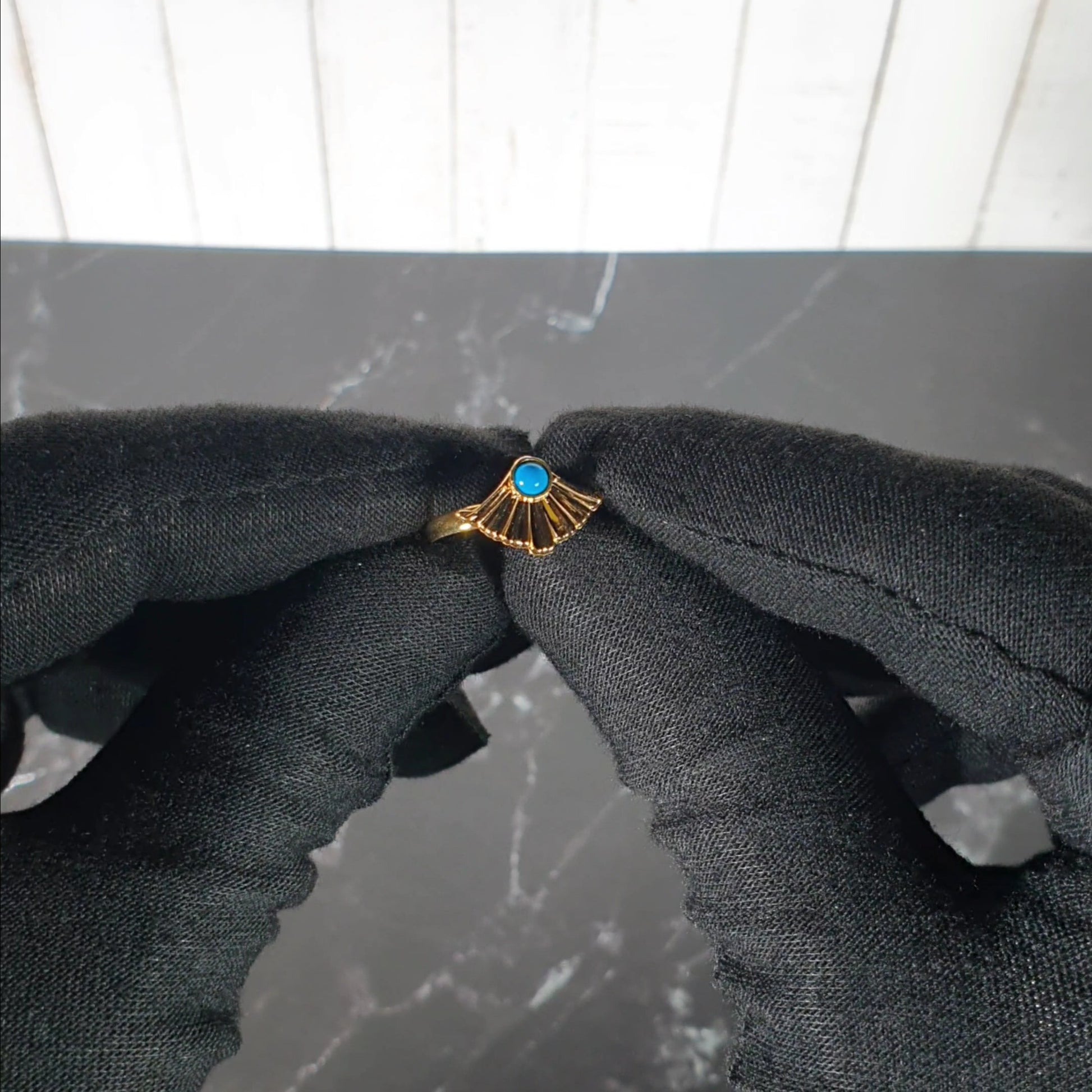 Gold ring with a blue gemstone held between black gloves on a gray background